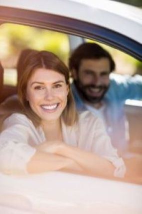 Hombre con traje sentado en el asiento del conductor de un automóvil, mirando a la cámara con una mujer rubia sonriendo a su lado.